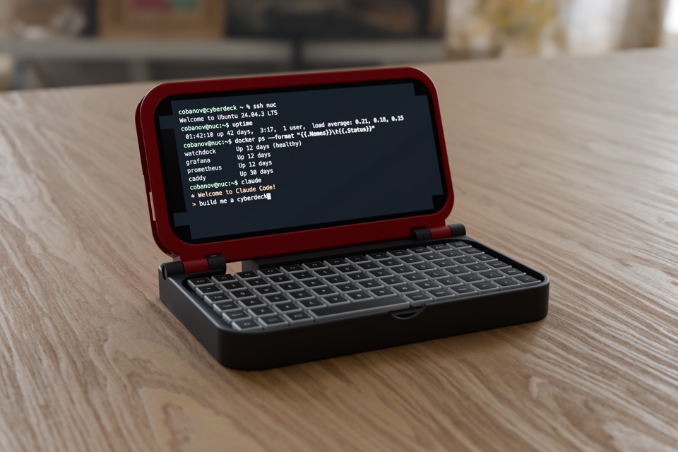 A red and black clamshell deck, open on a wooden table: an iPhone in the lid shows a terminal, a compact keyboard sits in the base.
